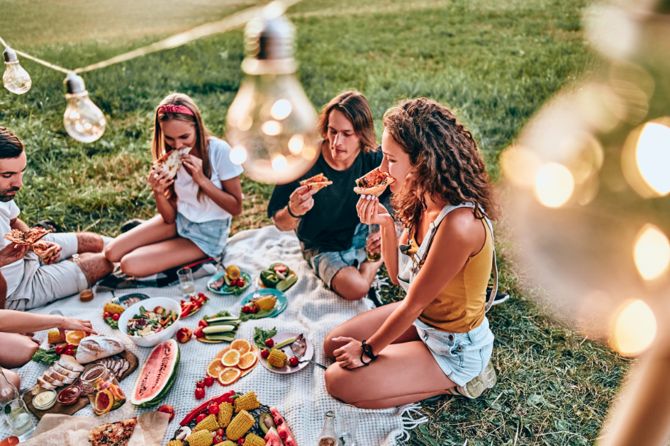 A group of friends gathers for a picnic of fresh fruits and savory foods under string lights.