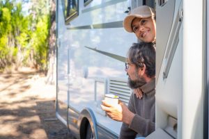 Couple leans out of their parked RV door with coffee in their hands on a beautiful summer morning