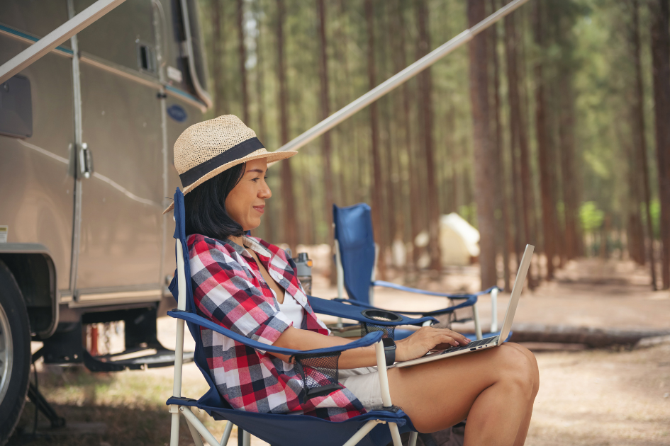 Woman sits outside her parked travel trailer with her laptop on her lap