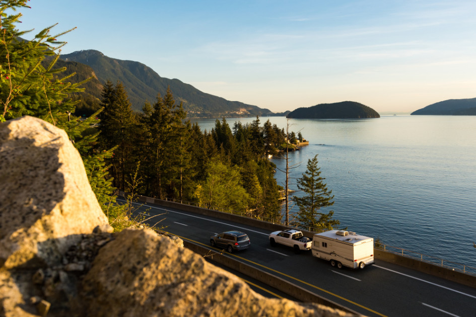 Truck towing a travel trailer behind it, driving down a scenic road next to a body of water with mountains in the distance